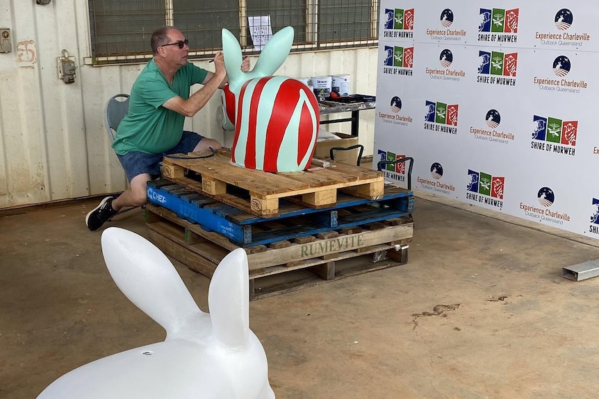 a man paints a cement bilby on a wooden pallet, with an unpainted bilby in the foreground