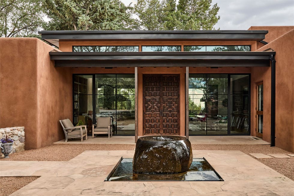 Modern adobe-style house exterior with a water feature and wooden door surrounded by greenery.