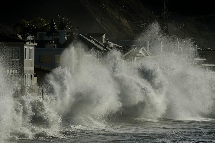 Huge waves crash up against a coastline. Their spray goes as high as the homes that are right next to the water.