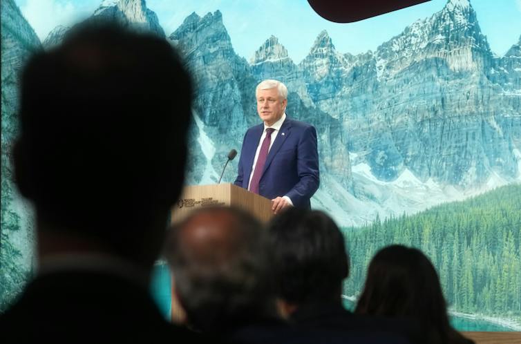 Older white man in a suit speaks from behind a podium to a crowd of people