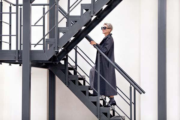 A woman in an ankle-length coat and large sunglasses climbs an exterior metal staircase.