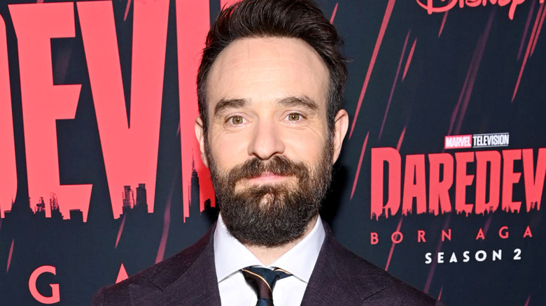 A man with short brown hair and medium-length brown beard smiles for the camera. He's wearing a dark purple suit and is stood against a red and black background with the logo for "Daredevil: Born Again" Season 2 on in red text.