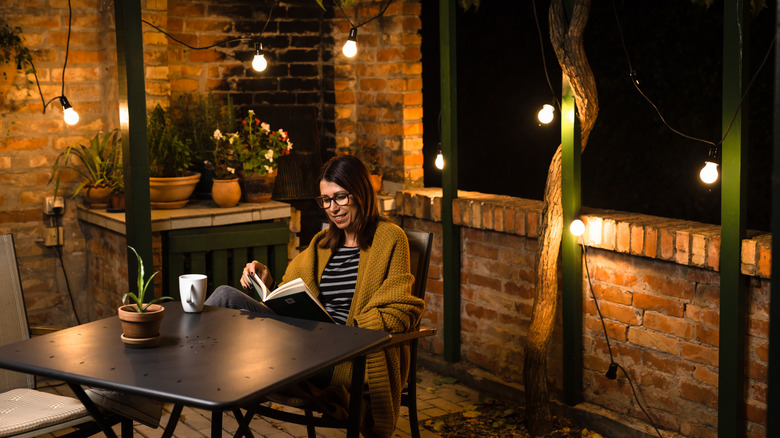 Person in a sweater, reading a book on a brick balcony lit with string lights