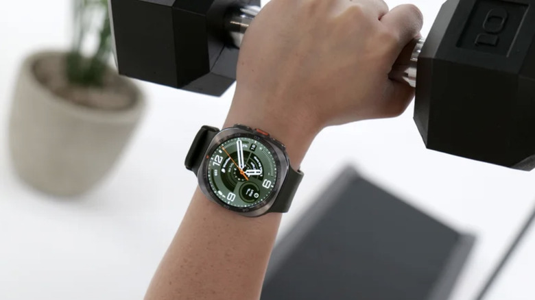 Close-up photo of a Samsung Galaxy Watch Ultra 2 smartphone on a wrist. The wearer is holding a black dumbbell above a blurred white background.