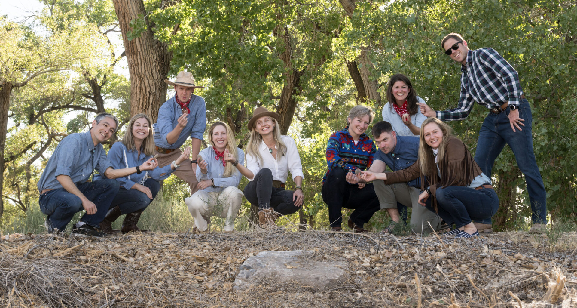 A group of 10 men and women wearing assorted jeans and button-down shirts hold up rock samples, with a forest in the background.