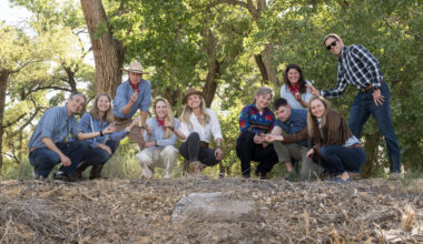 A group of 10 men and women wearing assorted jeans and button-down shirts hold up rock samples, with a forest in the background.