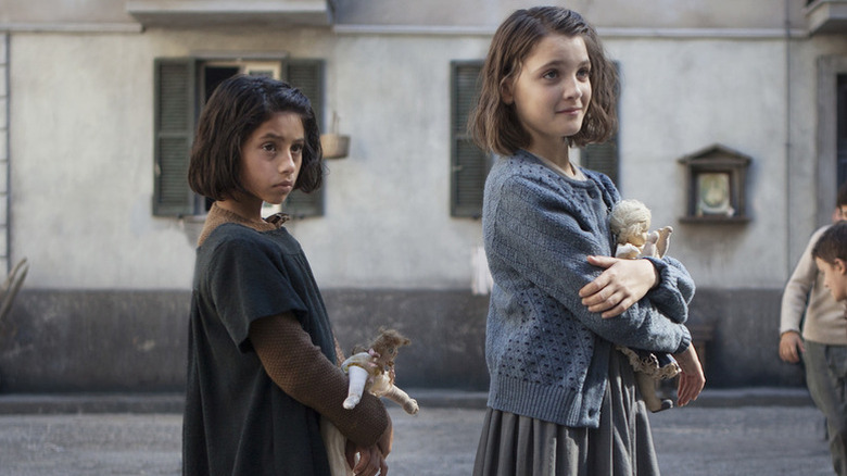 Lila holding doll next to Lenù, also holding doll, on the street