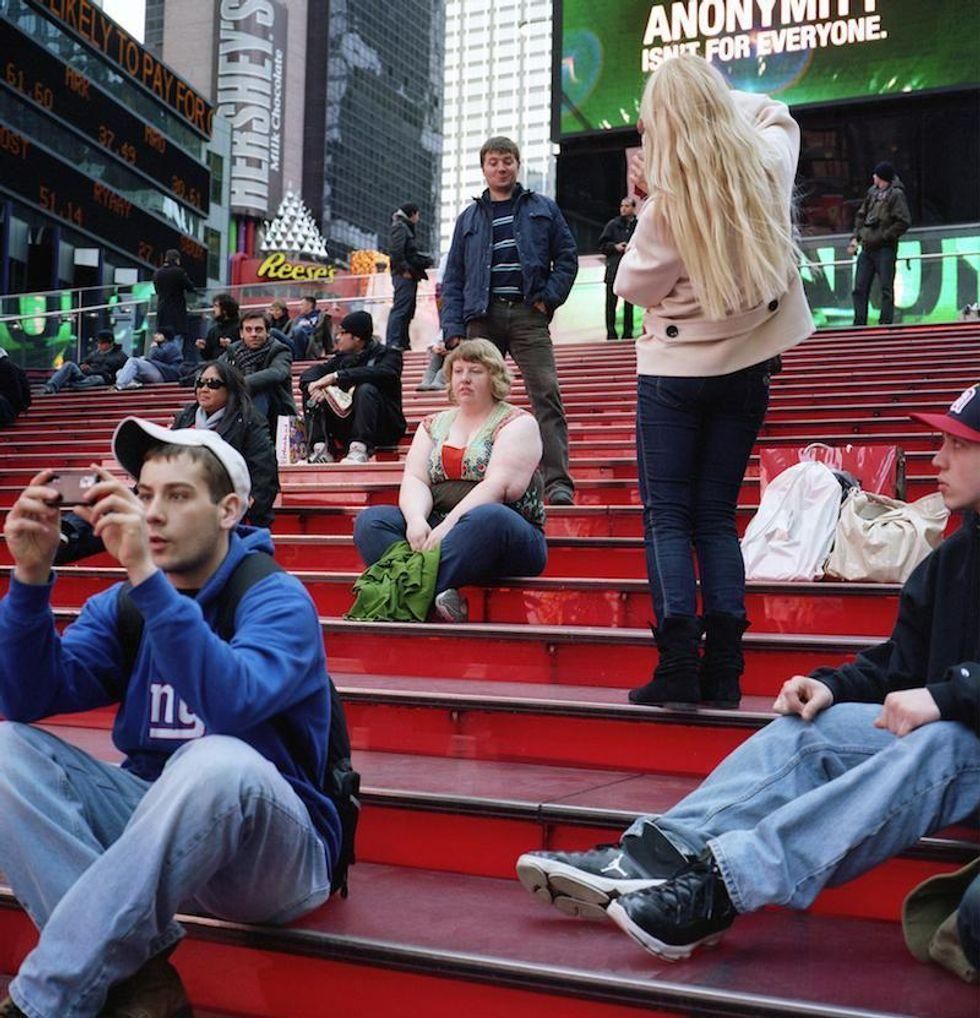 A girl standing in the bleachers of a sporting event taking a picture of her male friend, who is standing behind and above a heavy set woman, making a funny face as if to mock her.