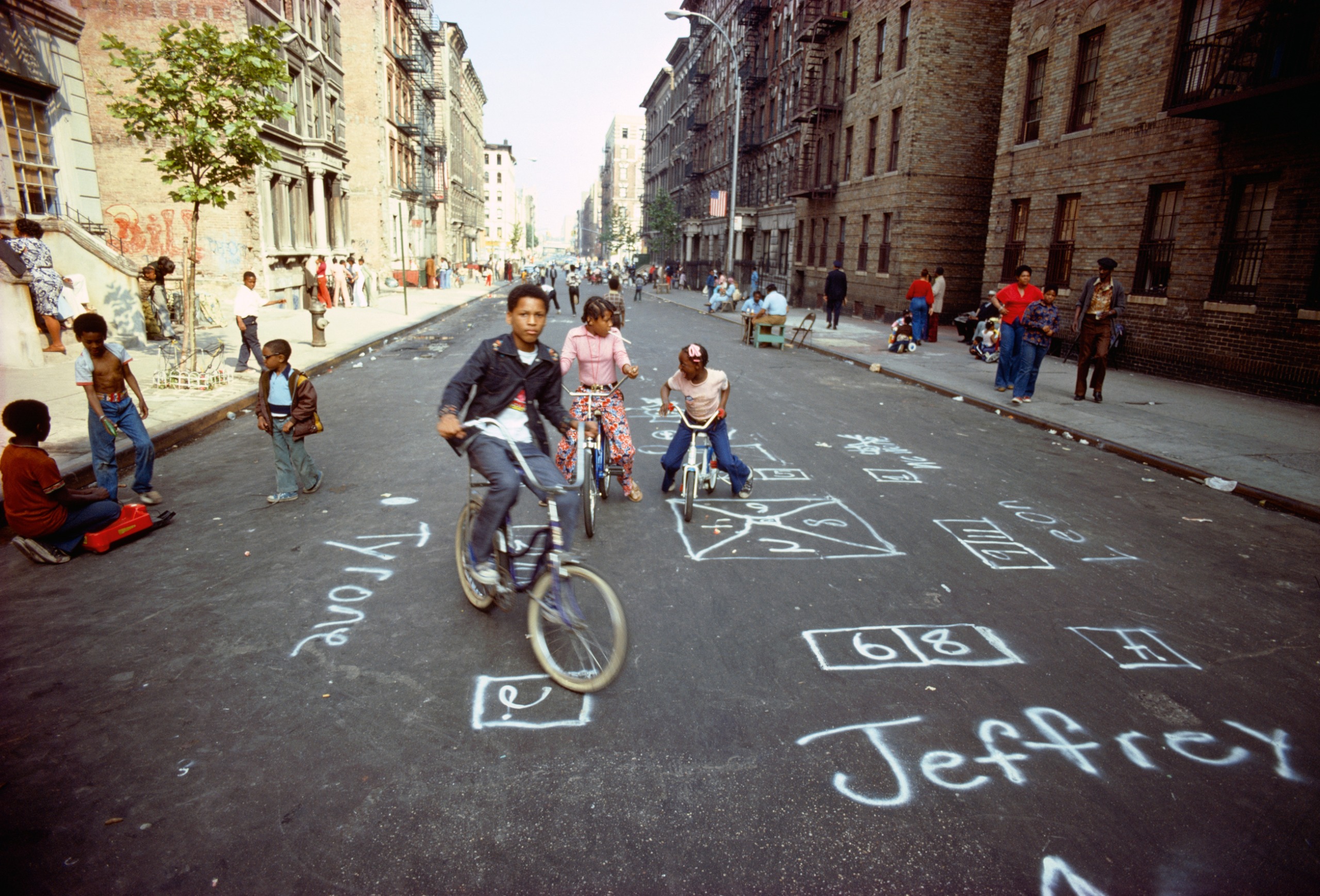 Children ride bicycles down a Harlem street chalked with hopscotch grids and names, flanked by brownstone apartment buildings.