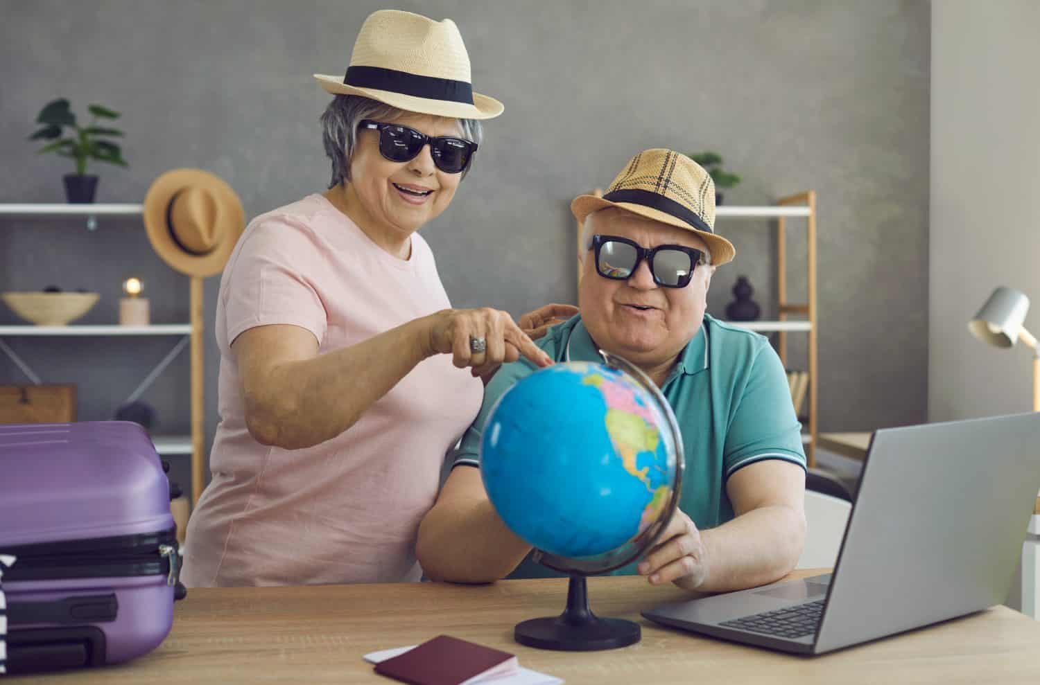 A smiling older woman in a pink shirt, straw hat, and sunglasses points at a blue globe while an older man in a green polo shirt, straw hat, and sunglasses holds the globe. A purple suitcase and passports are visible on a wooden desk with a laptop in a home setting.