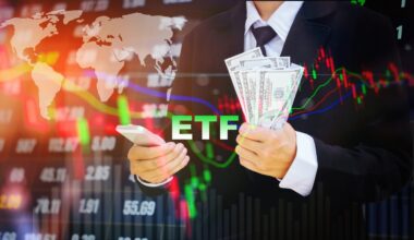 A person in a dark suit holds a silver smartphone in their left hand and a fan of one hundred U.S. dollar bills in their right hand. The green letters 'ETF' are visible in the center foreground. The background is a blurred, glowing digital display showing financial market data, including a world map, stock charts with red and green candlesticks, and various numerical figures, all overlaid with colorful trend lines.