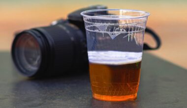 A clear plastic cup filled with beer sits on a dark surface in front of a blurred camera.