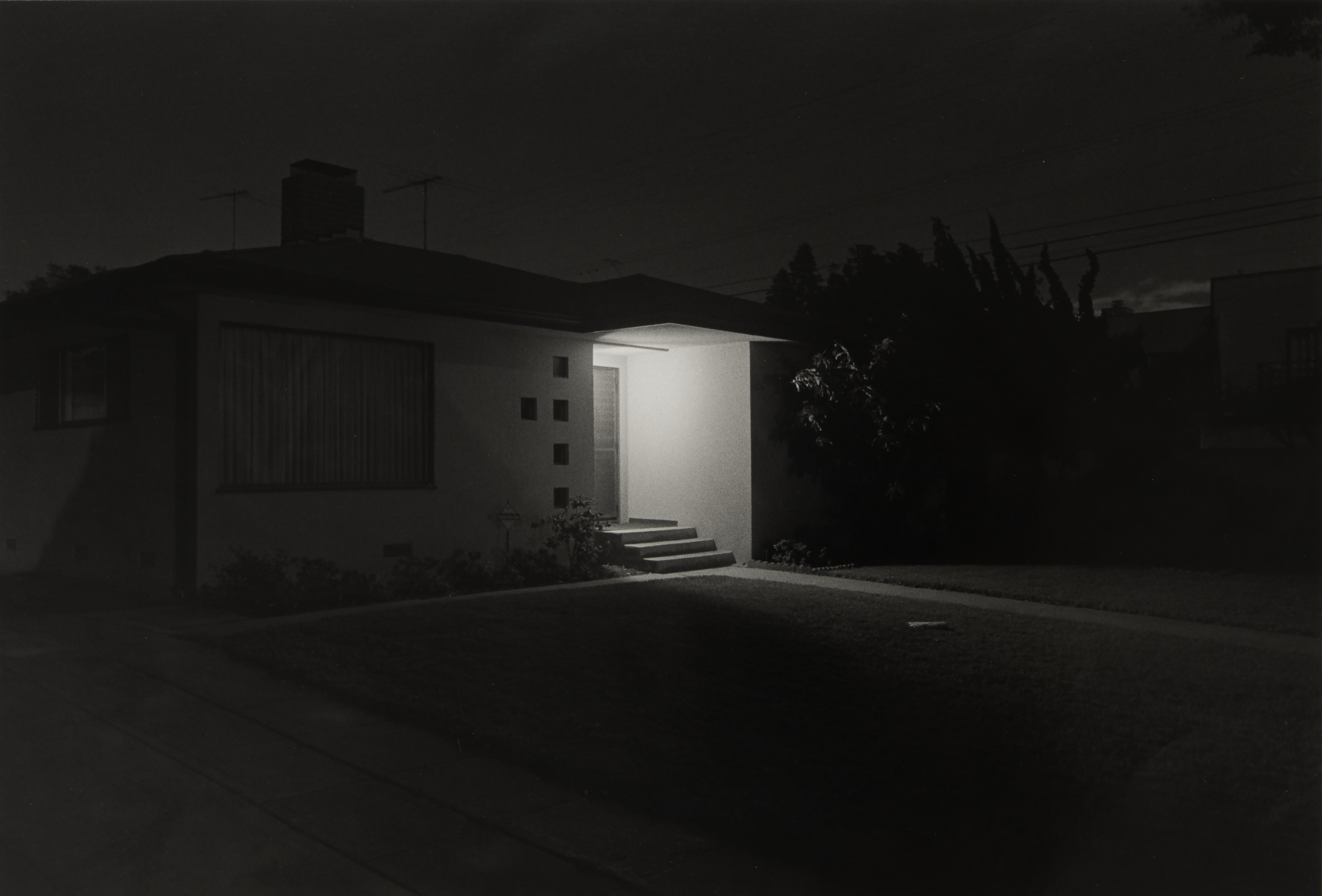 A black and white photograph of a house at night, its entryway lit by a single bright light against dark surroundings and trees.