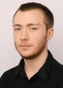 A young man with short brown hair and a beard is wearing a black collared shirt. He is looking slightly to the side with a neutral expression. The background is plain and light-colored.