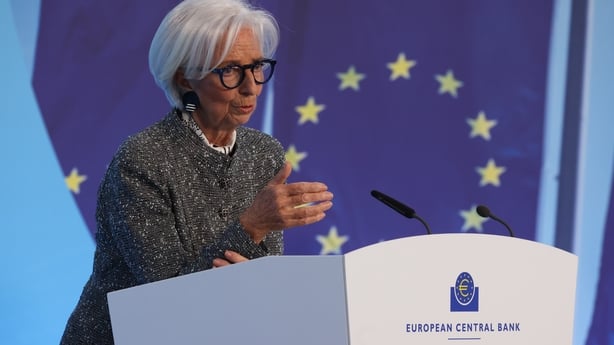 A woman in a grey suit speaks at a podium with the European Union flag in the background