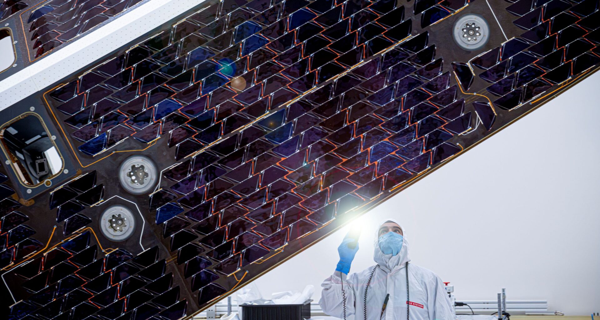 A technician in a white clean room gown and face mask shines a flashlight to inspect the dark, patterned cells of a large spacecraft solar array inside a clean room.