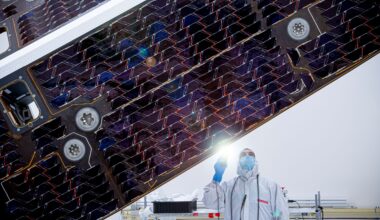 A technician in a white clean room gown and face mask shines a flashlight to inspect the dark, patterned cells of a large spacecraft solar array inside a clean room.