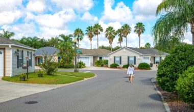 An overhead view of a well-maintained residential street in a warm climate, with single-story beige houses featuring dark shutters and attached garages. Lush green lawns and numerous palm trees line the street, which curves gently to the right. A person in a white top and shorts walks away from the viewer down the asphalt street. The sky is bright blue with scattered white clouds.