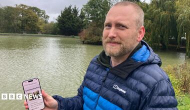 A man in a navy and light blue puffer jacket holds up a phone. He is standing in front of a body of water.