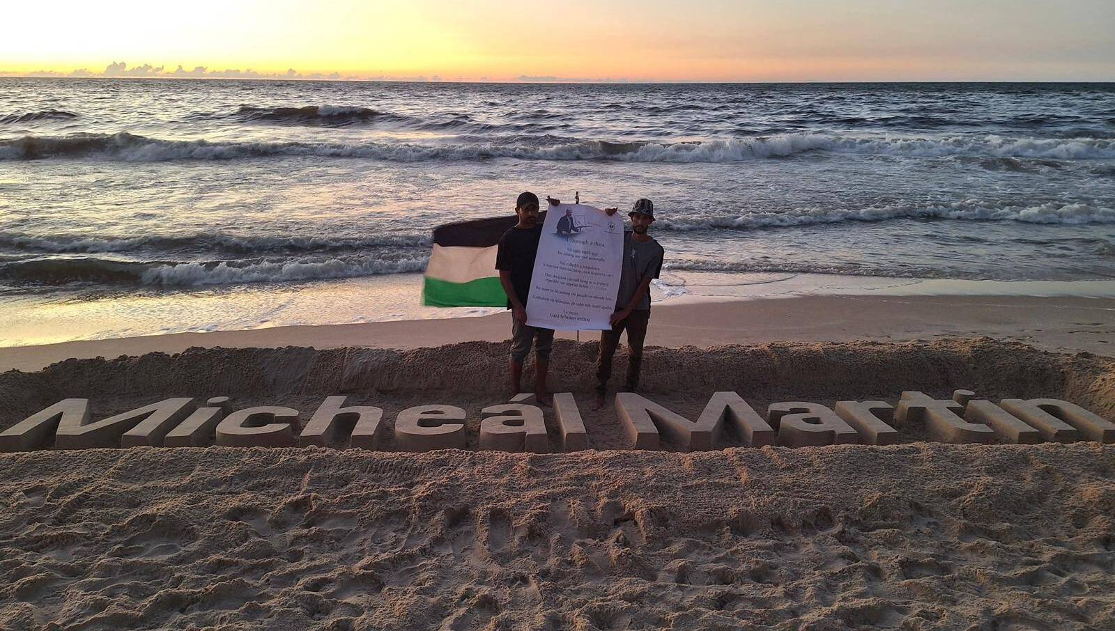 Two Gazan students refused study visas in Ireland pose beside a sculpture of Taoiseach Micheál Martin's name, carved in the sand at Gaza.