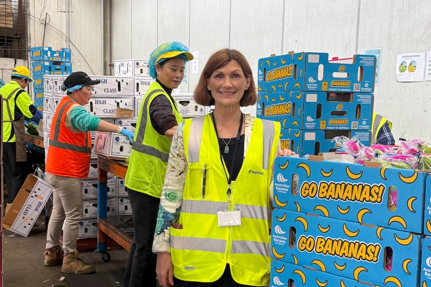 A woman in a yellow high-vis vest stands in a packing room, smiling beside stacked blue banana boxes.