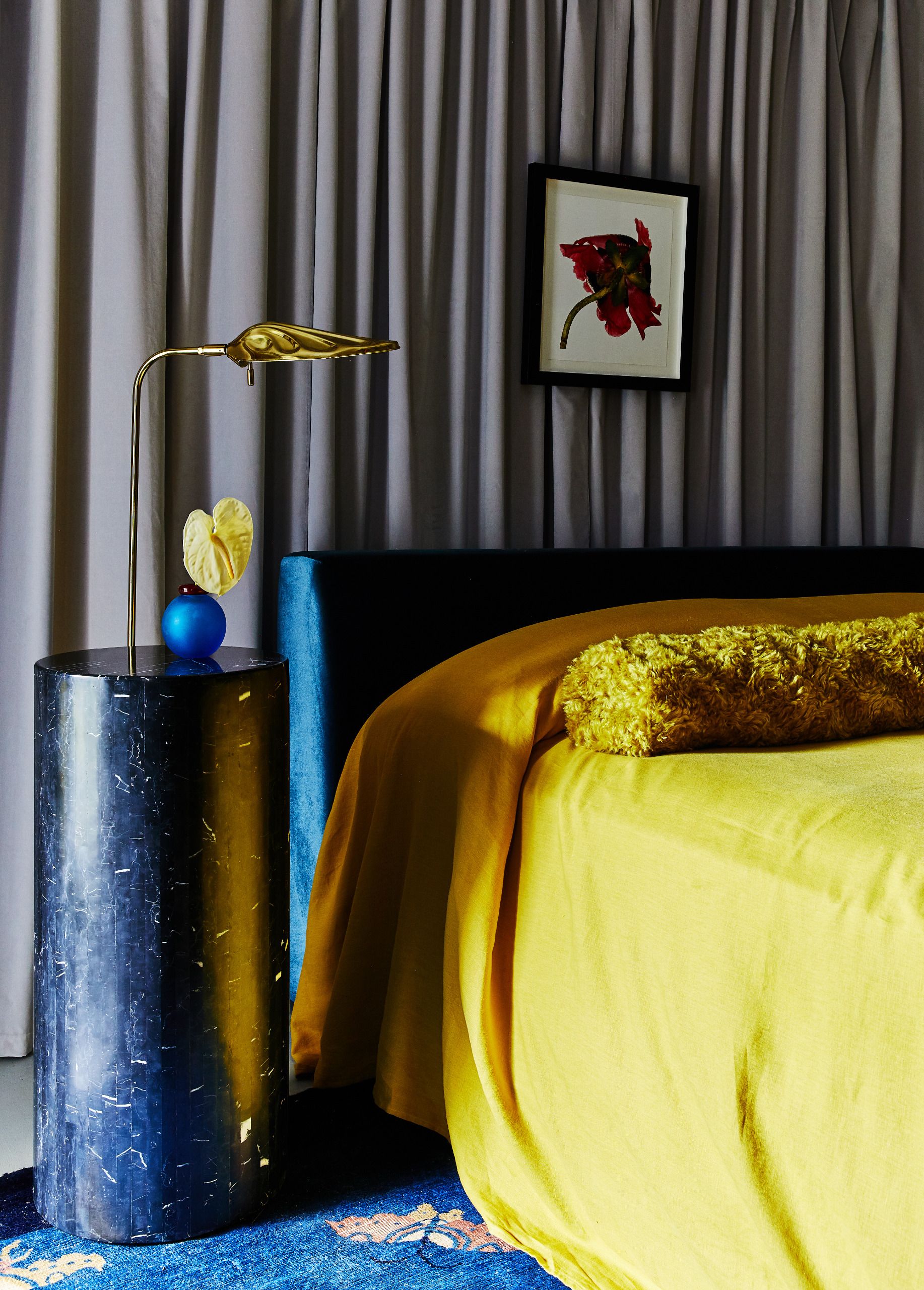 A bedroom with black marble pedestal table, bright ochre bedding and grey velvet curtain