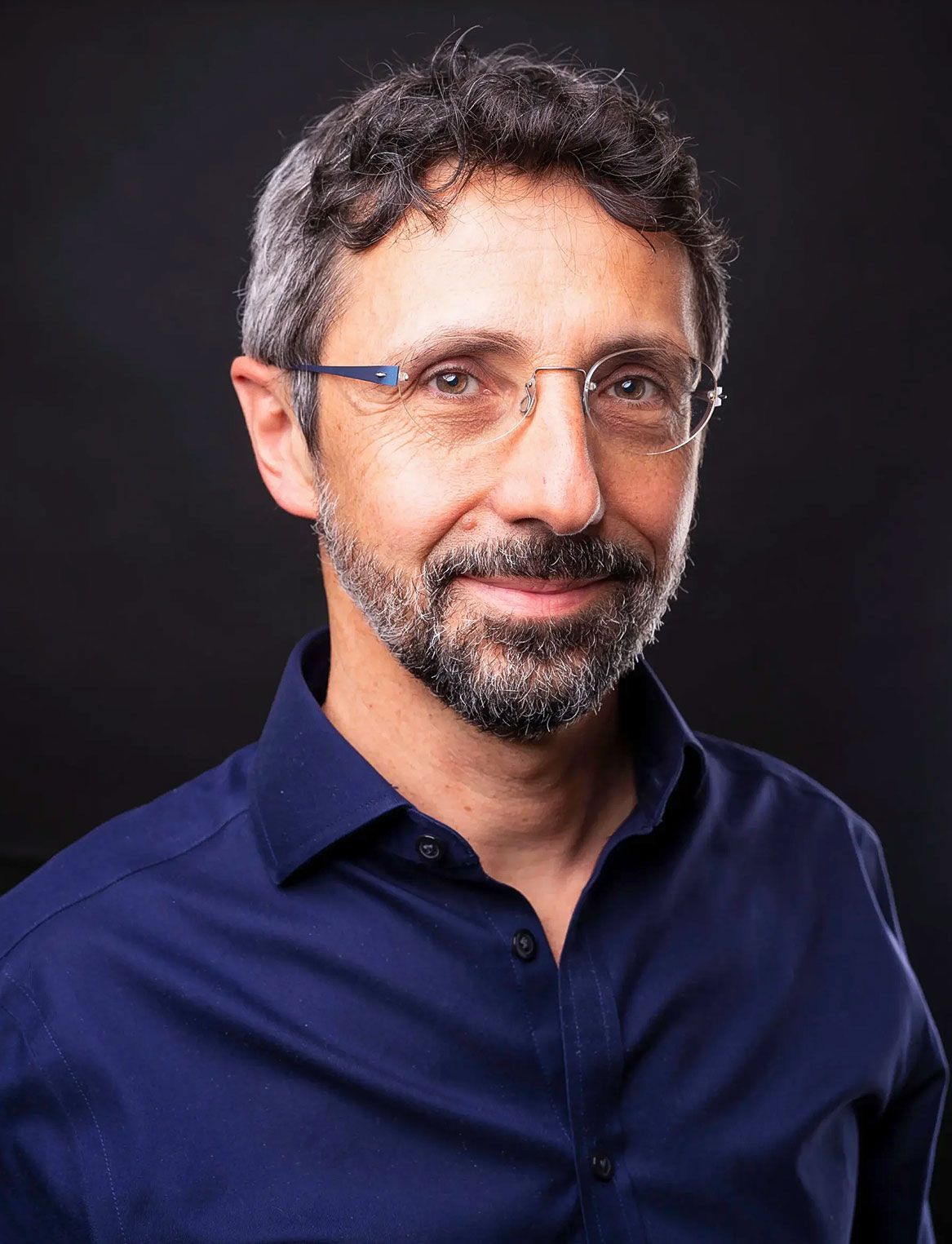 Headshot of a man with graying hair and a black beard with bits of gray. He wears rimless glasses and a dark blue button...