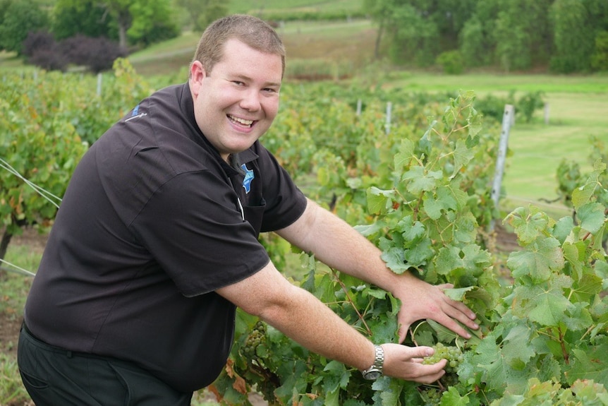 A man smiles next to a row of grapevines, reaching forward to display a bunch of green grapes.