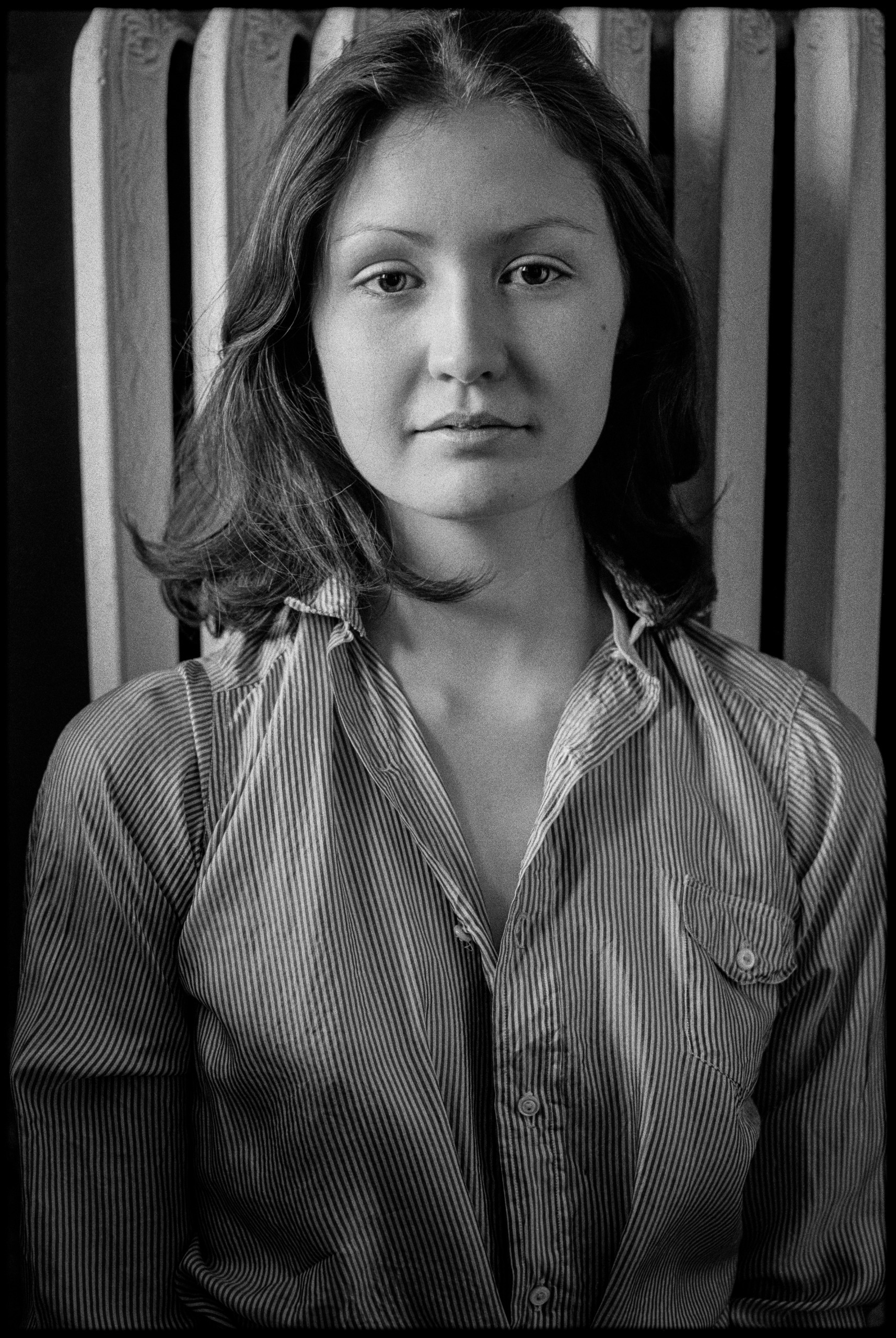 A black-and-white portrait of a young woman with shoulder-length wavy hair wearing a button-down striped shirt, posed in front of a radiator.