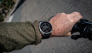 A cyclist's wrist in an olive fleece sleeve wearing a Garmin fenix watch with a silver bezel, its face showing 10:01, while the hand grips a bike handlebar on an asphalt road.