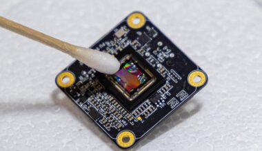 A black square with printed circuitry, tilted at an angle, against a white background. A wet q-tip points at the center of the square.