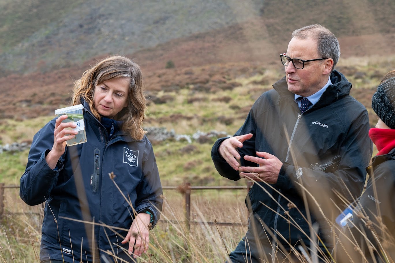 Helen Taylor holding a container of pond mud snails and Scott Arthur MP speaking in a natural habitat.