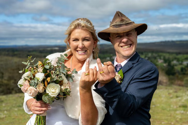 A smiling bride and groom showing their gold wedding rings to the camera.