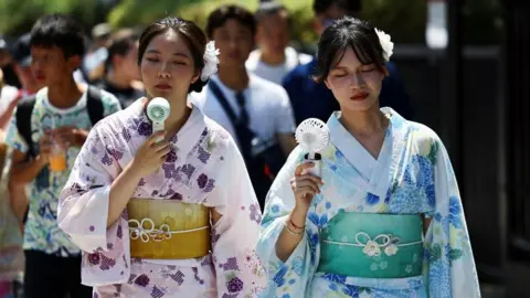 Reuters Two women use hand-held fans to cool themselves down. They both have dark hair; the woman on the left is dressed in pink and the woman on the right in blue.