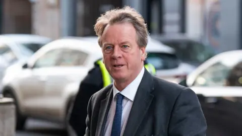 PA Media Alister Jack is wearing a dark suit jacket over a white shirt and a patterned blue tie. He has light brown/greyish hair that appears slightly tousled. The background features parked cars.