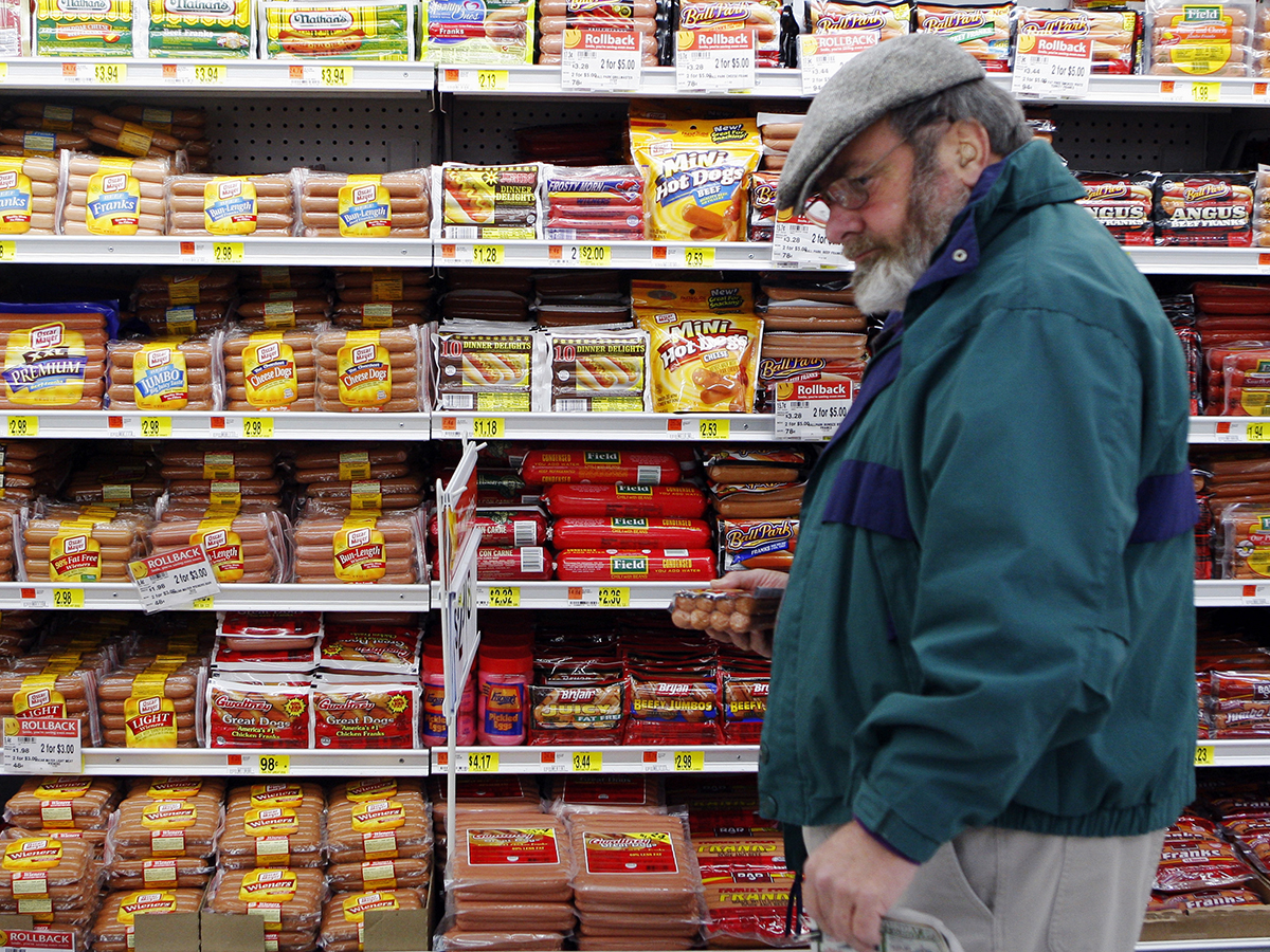 A bearded middle-aged man wearing glasses and a tam-style hat picks up a package of wieners in a WalMart grocery store.