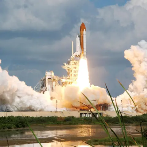 NASA Space Shuttle during launch at Cape Canaveral in Florida. A white shuttle is attached vertically to an orange rocket with large white booster engines. Flames are firing out of the rocket engine and there re large amounts of white smoke as the rocket is just about to lift off. In the foreground there is a a lake.