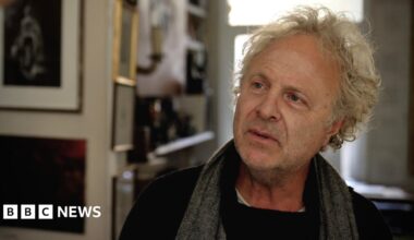 A tree branch with red leaves in the foreground, with smaller trees planted in plastic protective supports in a meadow backed by mature trees and a sunlit, mown field beneath a partially cloudy sky