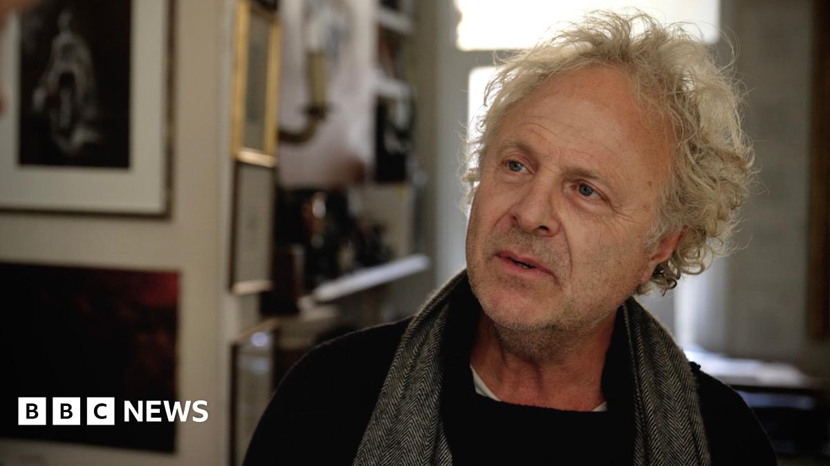 A tree branch with red leaves in the foreground, with smaller trees planted in plastic protective supports in a meadow backed by mature trees and a sunlit, mown field beneath a partially cloudy sky