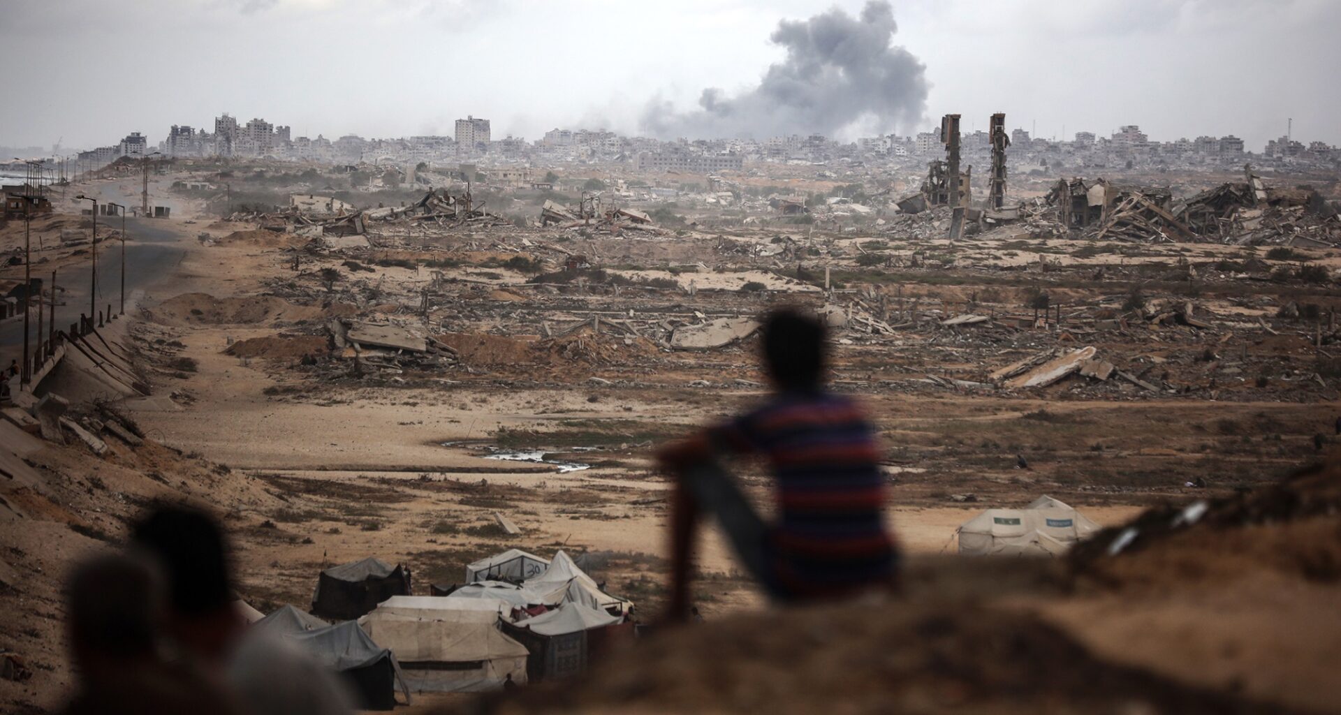 Two figures are backlit by lights from a car as they walk down a street. Other people are visible at the edges of the frame.