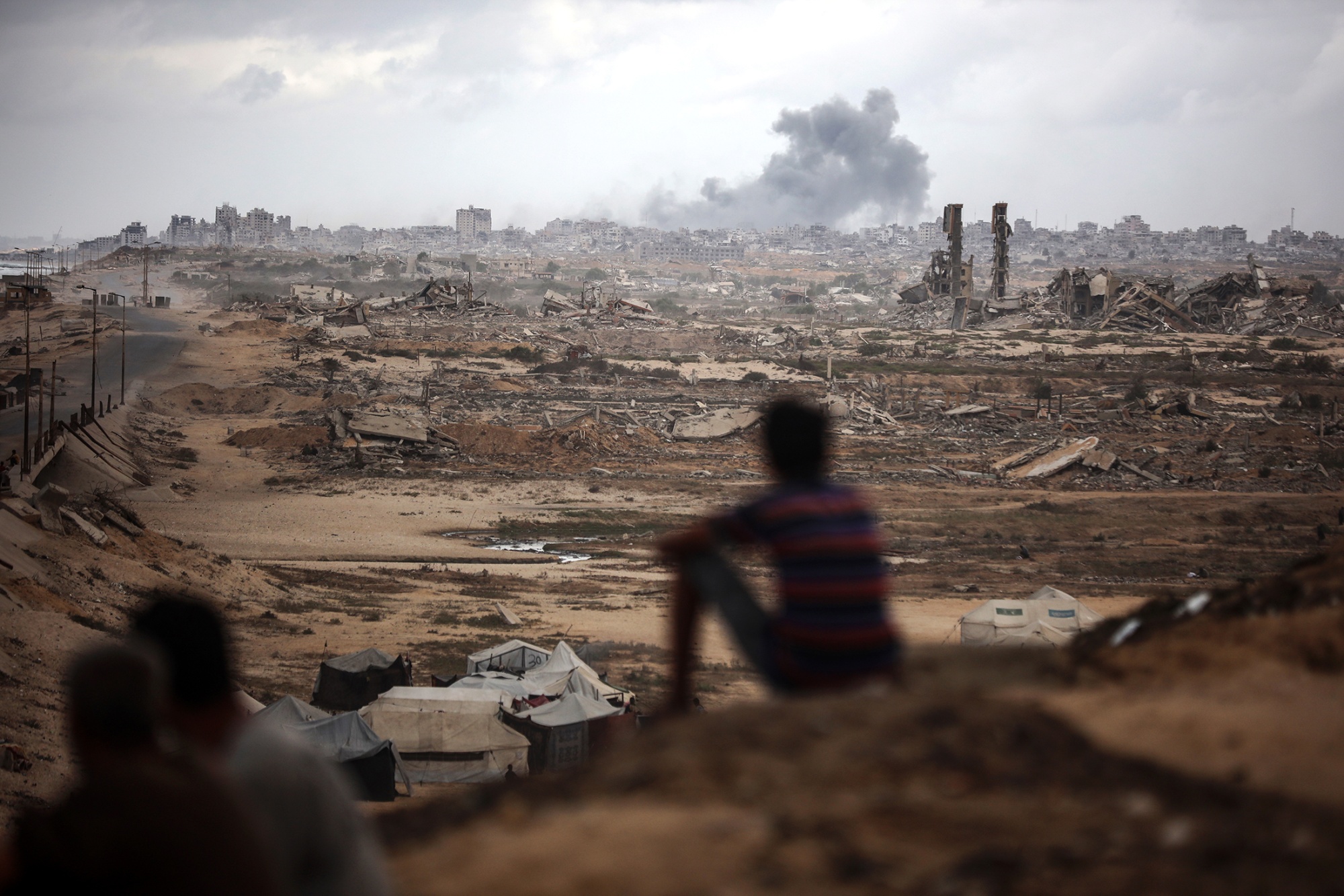 People sit on a hill looking out at a plume of smoke over ruined buildings on the horizon.