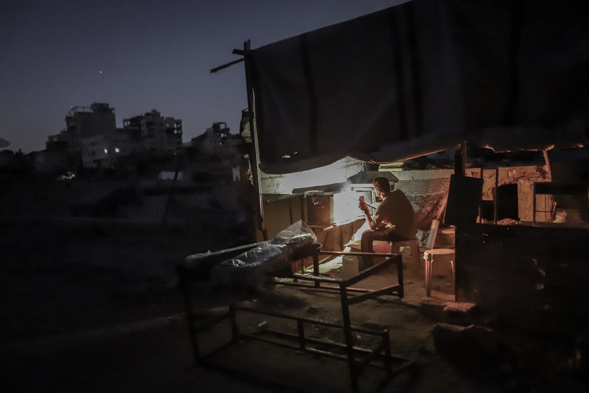 A person lights a fire in a makeshift tent structure against a dark sky.