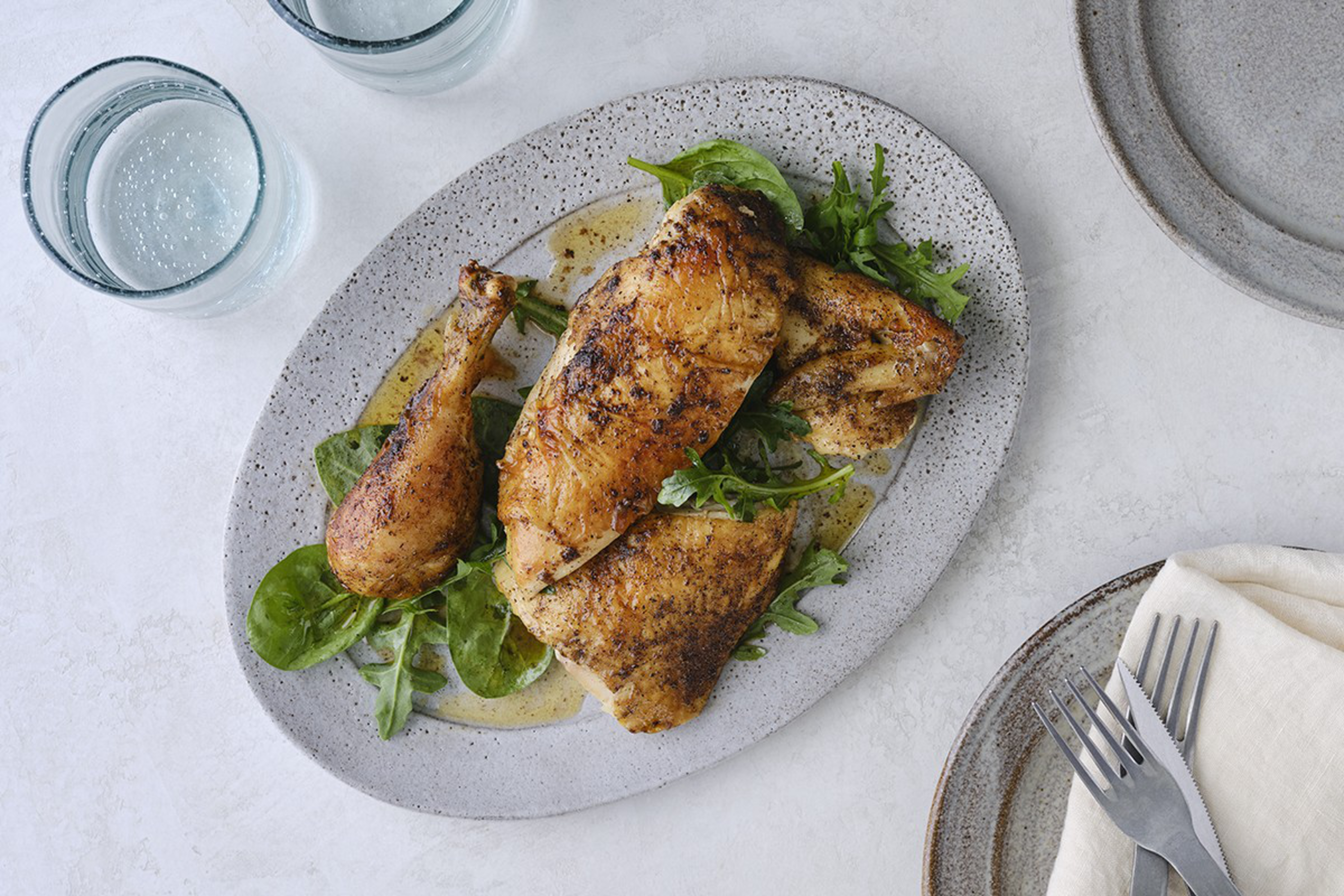A speckled platter holds seasoned roasted chicken pieces on fresh greens, beside two glasses of water and empty stoneware plates with cutlery wrapped in a napkin.