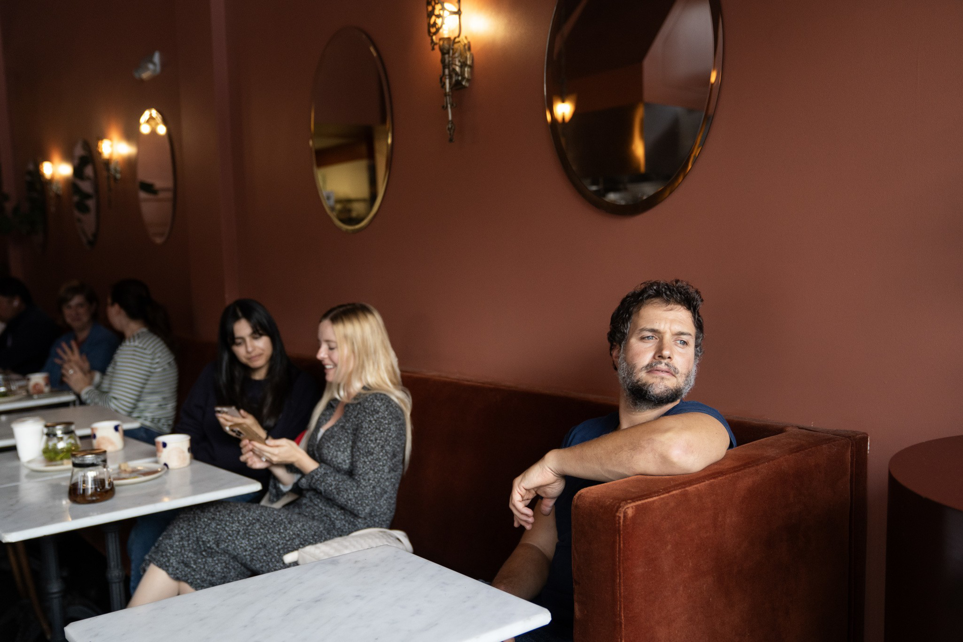 A man sits alone on a red velvet bench looking away while two women nearby share a phone, with round mirrors and warm lights on a brown wall behind them.