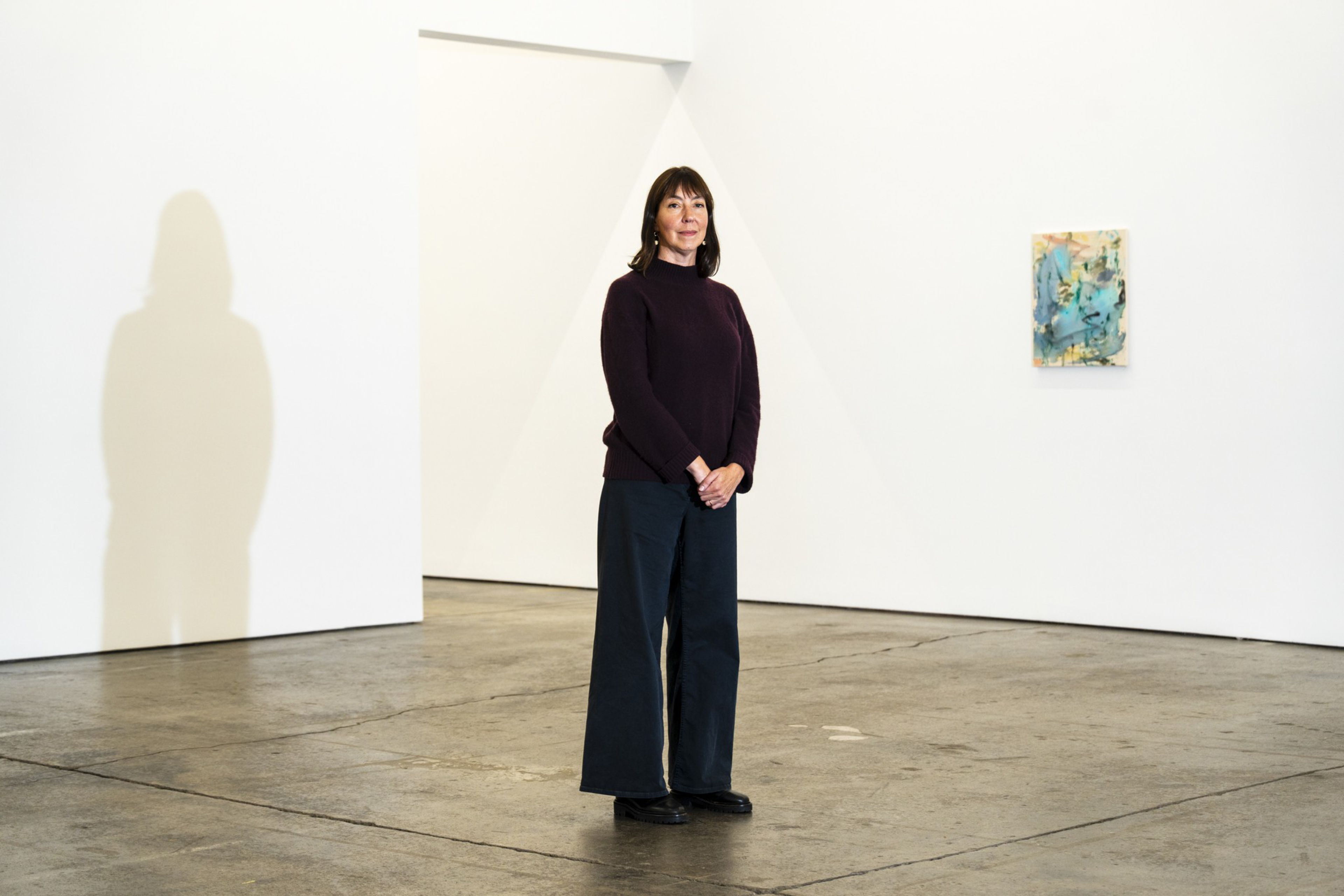 A woman stands with hands clasped in an empty gallery with concrete floors, wearing a dark sweater and wide-leg pants, with a small colorful painting on the wall.