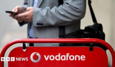 A man wearing a suit and holding his phone stands in front of a Vodafone sign.