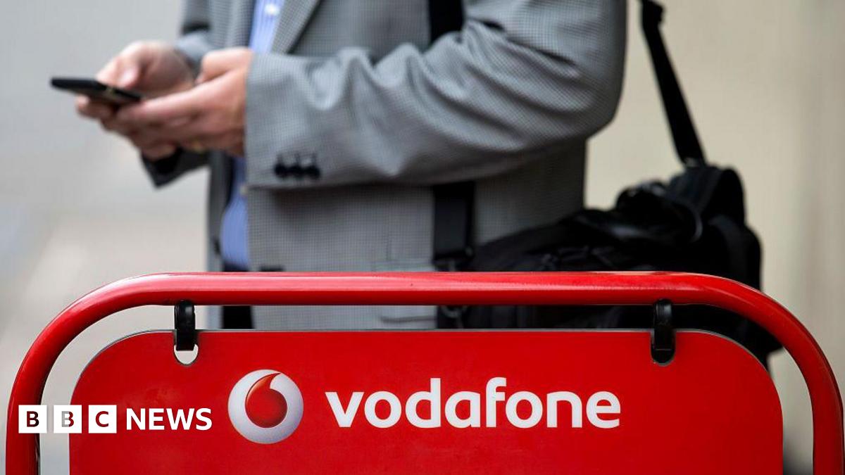 A man wearing a suit and holding his phone stands in front of a Vodafone sign.