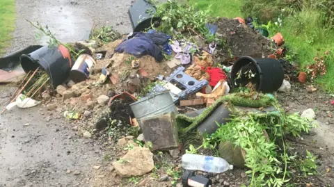 Huw Griffiths Rubbish piled together on a country road. There are black plastic and grey metal buckets, plastic bottles, garden waste and household items. 