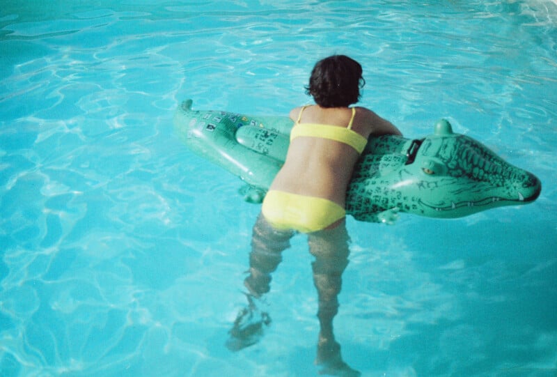 A child in a yellow swimsuit floats on a green inflatable alligator in a swimming pool with clear blue water.
