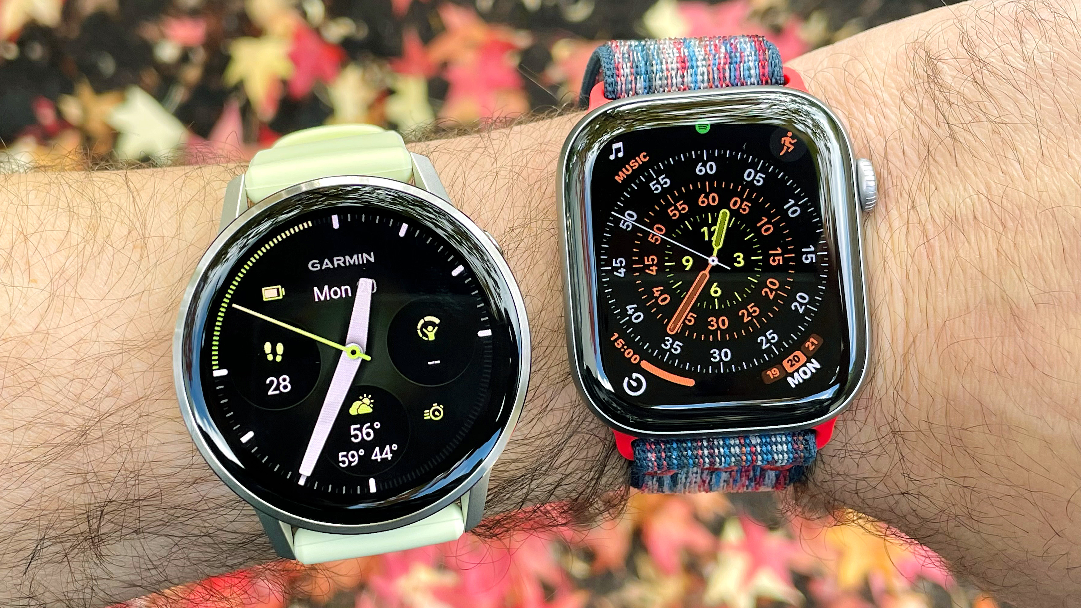 Close-up of the Garmin Venu 4 next to the Apple Watch Series 11 on a user's wrist with foliage out of focus in the background